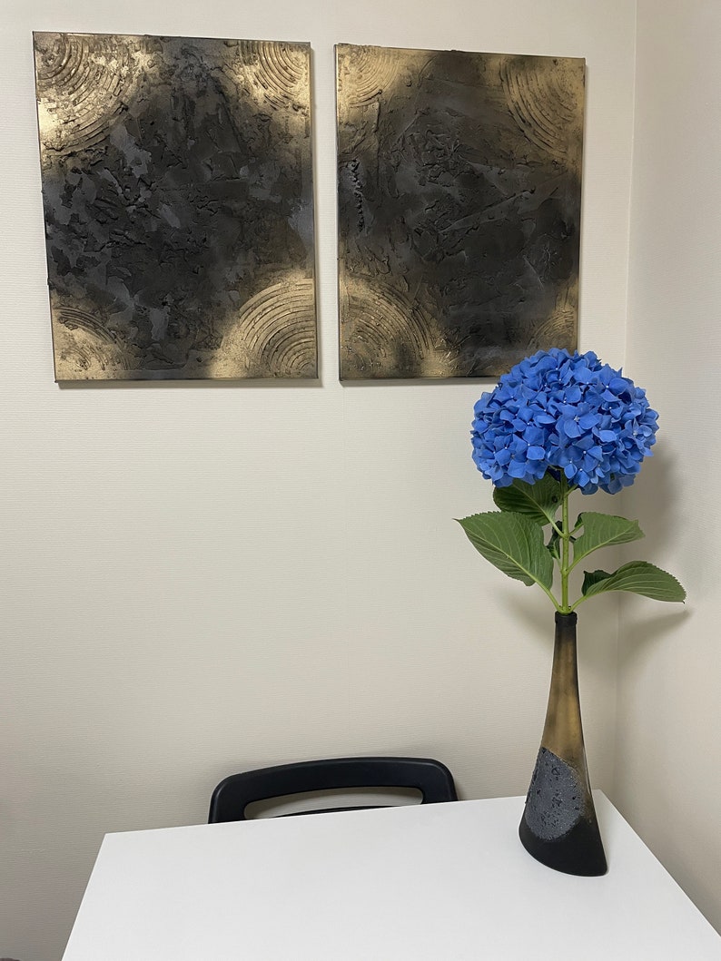 May include: Two abstract paintings with black and gold textured paint on canvas, hanging on a white wall. A blue hydrangea flower in a black and gold vase sits on a white table with a black chair.