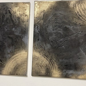 May include: Two square abstract paintings with a textured black and gold design. The paintings are side by side on a white wall.