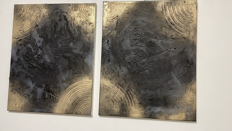 May include: Two square abstract paintings with a textured black and gold design. The paintings are side by side on a white wall.