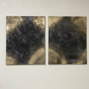 May include: Two square abstract paintings with a black and gold color scheme. The paintings have a textured surface and a circular pattern.
