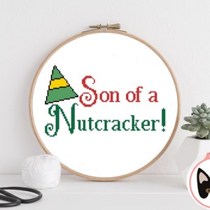 May include: A white embroidery hoop with a green and yellow elf hat and the text "Son of a Nutcracker!" in red and green lettering.
