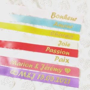 Op de afbeelding: Acht satijnen linten in verschillende kleuren met gouden glitterletters. De linten hebben de woorden "Bonheur", "Amour", "Cher", "Joie", "Passion", "Paix", "Marion & Jérémy" en "M&J 18.05.2021" erop geschreven.