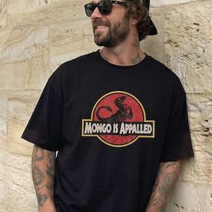 May include: A black short-sleeved t-shirt featuring a circular graphic with a red background and yellow border. The graphic depicts a black dinosaur silhouette and the text "MONGO IS APPALLED" in a bold, white, serif font across the centre.