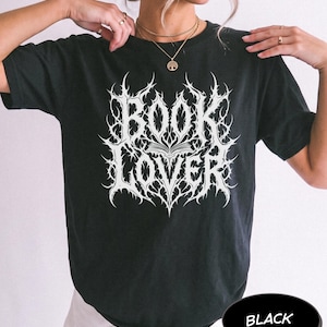 May include: A black short-sleeved t-shirt featuring a white graphic print on the chest. The design displays the text "BOOK LOVER" in a stylised, sharp, thorn-like font with an open book icon centred between the two words.