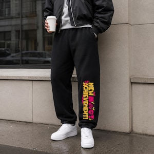 May include: Black sweatpants featuring a vertical graphic on the left leg with the text "NEW ACHIEVEMENT!" in yellow, accompanied by pink icons including a cube, a lightning bolt, a skull, and a trophy, paired with white sneakers.