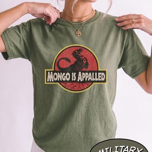 May include: A military green short-sleeve t-shirt featuring a circular graphic with a red background and a black dinosaur silhouette. The text "MONGO IS APPALED" is printed in white capital letters inside a yellow-bordered black rectangular banner.