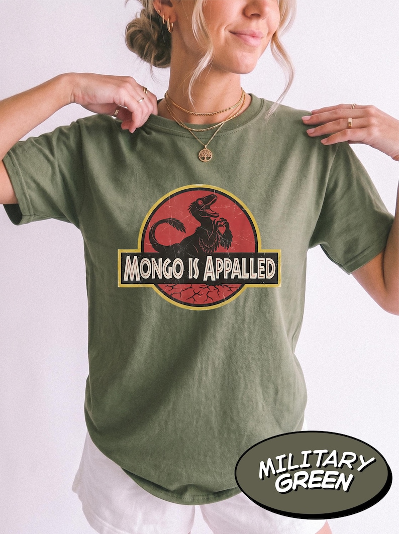 May include: A military green short-sleeve t-shirt featuring a circular graphic with a red background and a black dinosaur silhouette. The text "MONGO IS APPALED" is printed in white capital letters inside a yellow-bordered black rectangular banner.