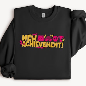 May include: A black long-sleeve sweatshirt featuring the yellow text NEW ACHIEVEMENT! with a pink graphic design including a pair of shorts, sunglasses, a skull, and a trophy, accented by pink lightning bolt shapes on the sides of the text.