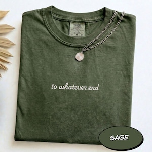Pode incluir: Uma t-shirt de manga curta verde sálvia com decote redondo e o texto branco bordado "to whatever end" centrado no peito. Um colar de corrente prateada com um pendente redondo repousa sobre a gola. A t-shirt tem uma etiqueta Comfort Colors.