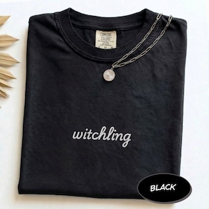 May include: A black short-sleeved t-shirt featuring the word witchling embroidered in white script across the chest. The shirt has a Comfort Colors brand label at the inner neckline and is paired with a silver-toned chain necklace with a round pendant.