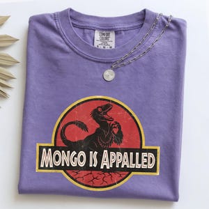 May include: A purple t-shirt featuring a graphic of a black dinosaur inside a red circle with a yellow border. The text "MONGO IS APPALED" is printed in white capital letters on a black rectangular banner. A silver chain necklace rests on the collar.