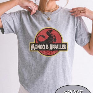 May include: A sport grey t-shirt featuring a circular graphic with a black dinosaur silhouette on a red background. A yellow banner across the center contains the black text "MONGO IS APPALED" in a bold, sans-serif font. The design has a distressed texture.