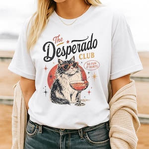 Puede incluir: Una camiseta blanca con un gráfico de un gato esponjoso con gafas de sol rojas junto a una copa de cóctel. El texto dice The Desperado CLUB y SO FUN IT HURTS, acompañado de iconos de estrellas y un elemento circular rojo tras el gato.