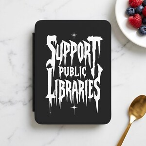 May include: A black rectangular e-reader case featuring white text that reads SUPPORT PUBLIC LIBRARIES in a stylised, dripping font. The design includes two small four-pointed star icons positioned above and below the central text.