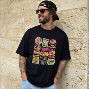 May include: A black short-sleeved t-shirt featuring a colourful graphic print with the text Dungeon Crawler Carl, surrounded by various small illustrations and phrases including God Damnit Donut, You Will Not Break Me, and New Achievement.