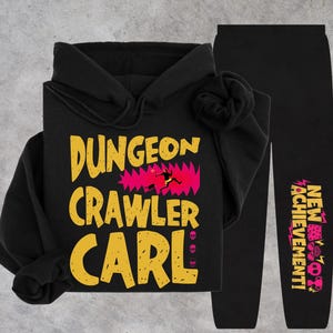 Może przedstawiać: Czarna bluza z kapturem i pasujące spodnie dresowe. Bluza posiada duży żółty napis DUNGEON CRAWLER CARL oraz różową grafikę. Na spodniach znajduje się żółto-różowy napis NEW ACHIEVEMENT umieszczony wzdłuż nogawki.