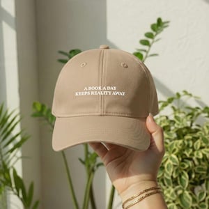 May include: A beige baseball cap featuring the white embroidered text A BOOK A DAY KEEPS REALITY AWAY centered on the front panel. The cap has a structured crown, a curved brim, and stitched eyelets on the top panels for ventilation.