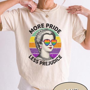 May include: A sand-colored t-shirt featuring a graphic of a historical figure wearing heart-shaped rainbow sunglasses. The design includes a circular background with horizontal stripes in yellow, orange, and purple, and the text MORE PRIDE LESS PREJUDICE.