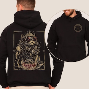 May include: A black hooded sweatshirt featuring a large graphic on the back of a long-haired cat wearing a crown and round sunglasses, sitting behind a bowl of cherries. The front chest area displays a circular logo with a dagger and text.