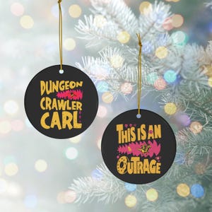 May include: Two round black ceramic ornaments with gold hanging strings. One ornament features the yellow text Dungeon Crawler Carl with a pink graphic. The other ornament displays the yellow text This Is An Outrage with a pink graphic.