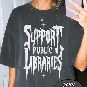 May include: A dark heather grey short-sleeve t-shirt featuring white graphic text that reads SUPPORT PUBLIC LIBRARIES in a stylised, dripping font. The design includes small four-pointed star accents above and below the text, centred on the chest.