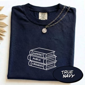 May include: A navy blue t-shirt featuring a white embroidered graphic of a stack of three books with the text "SUPPORT PUBLIC LIBRARIES" on the spines. A silver-toned chain necklace with a round pendant rests on the neckline of the shirt.