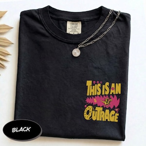 May include: A black short-sleeved t-shirt featuring a graphic on the lower right side with the yellow text "THIS IS AN OUTRAGE" above a pink jagged shape, a small dinosaur, and a doughnut. A silver chain necklace with a round pendant rests on the collar.