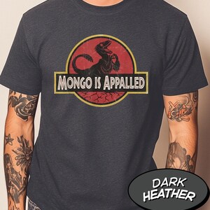 May include: A dark heather gray short-sleeve t-shirt featuring a circular graphic with a red background and a black dinosaur silhouette. The text "MONGO IS APPALED" is printed in white capital letters inside a yellow-bordered rectangular banner.