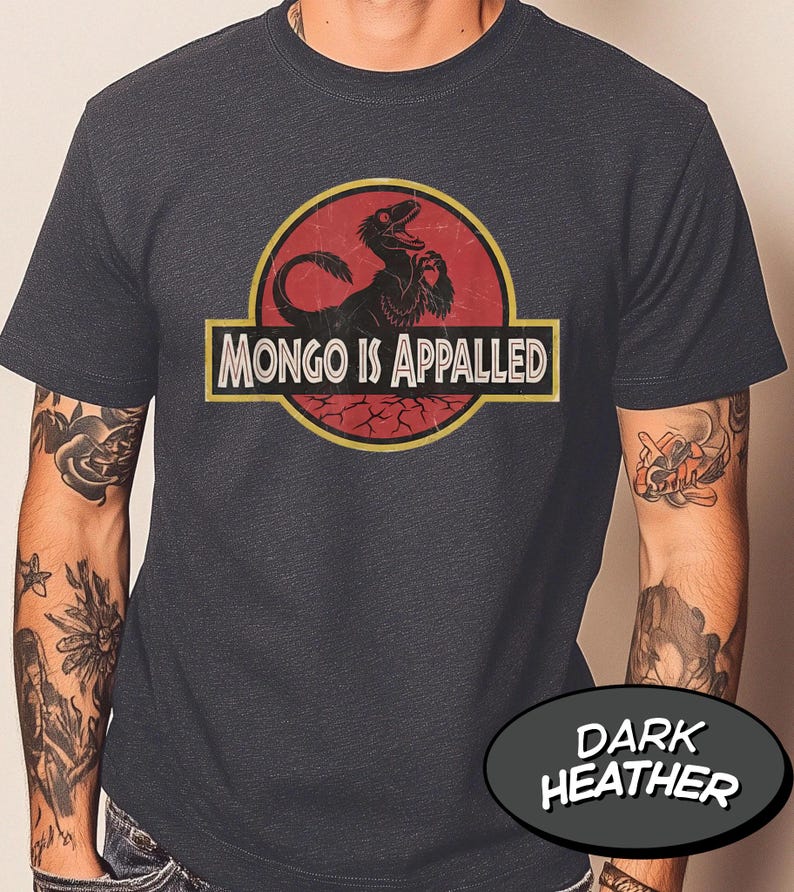 May include: A dark heather gray short-sleeve t-shirt featuring a circular graphic with a red background and a black dinosaur silhouette. The text "MONGO IS APPALED" is printed in white capital letters inside a yellow-bordered rectangular banner.