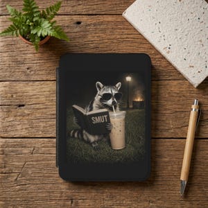May include: A black e-reader device displaying an illustration of a raccoon wearing sunglasses, holding a book titled SMUT, and drinking an iced coffee with a straw. The scene is set outdoors at night with a glowing lamp post in the background.