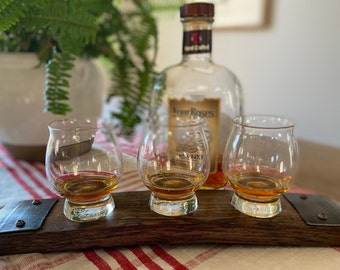 May include: Three clear whiskey glasses with amber liquid on a dark wooden serving board with metal details. A bottle of Four Roses Small Batch bourbon is in the background. The setup is on a red and white striped tablecloth.