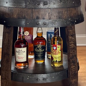 May include: A wooden barrel-shaped bar cart with a metal top and a metal bottom shelf. The cart is holding four bottles of liquor: Old Forester 1870 Original Batch, Weller 12 Year Old, Jeppson's Malort Liqueur, and Rabbit Hole Whiskey.