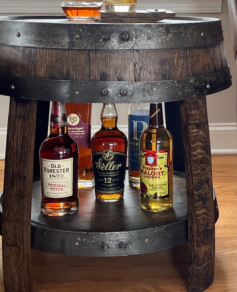 May include: A wooden barrel-shaped bar cart with a metal top and a metal bottom shelf. The cart is holding four bottles of liquor: Old Forester 1870 Original Batch, Weller 12 Year Old, Jeppson's Malort Liqueur, and Rabbit Hole Whiskey.