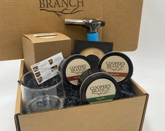 May include: A brown cardboard box with the words "Cooper's Branch" printed on the top. The box contains a glass, a small blue torch, a wooden coaster, and three small tins of wood chips labeled "Hickory", "Maple", and "Apple".
