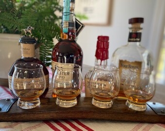 May include: A wooden barrel stave with four small glasses filled with amber liquid. The glasses are arranged in a row on the stave. There are four bottles of liquor behind the glasses, including Maker's Mark, Old Forester, and Wild Turkey.