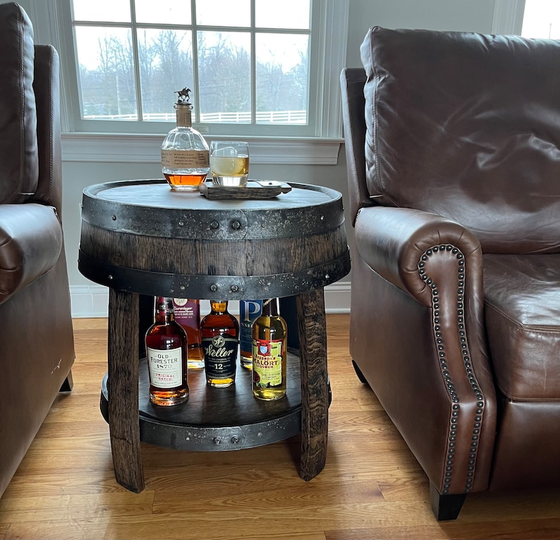 May include: A wooden barrel end table with a two-tiered shelf. The table has a dark brown finish and is decorated with metal accents. The top shelf is holding a glass of liquid and a bottle of liquor. The bottom shelf is holding four bottles of liquor. The table is sitting on a wooden floor.