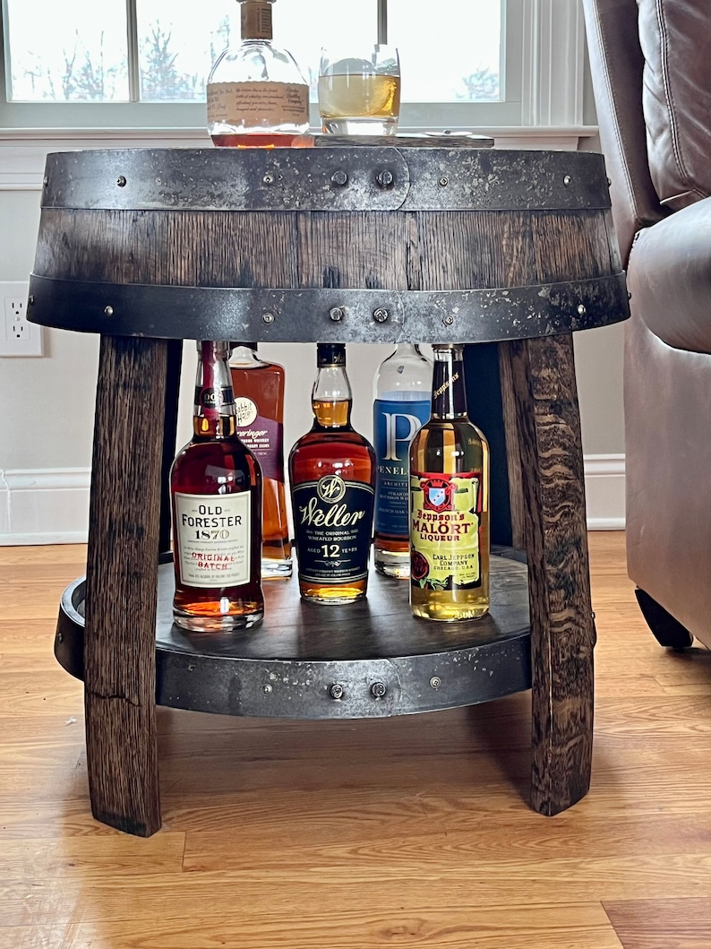 May include: A rustic wooden barrel table with a metal top and a lower shelf. The table is decorated with metal accents and has four legs. There are five bottles of liquor on the table, including Old Forester 1870, Weller 12 Year Old, and Jeppson's Mal&ouml;rt Liqueur.