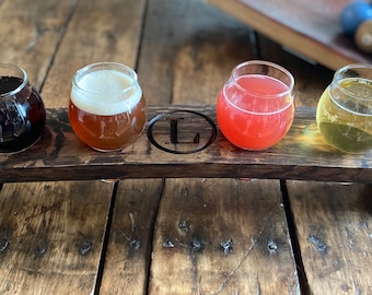 Puede incluir: Un tablero de vuelo de madera con cuatro vasos pequeños llenos de bebidas de diferentes colores. El tablero tiene un logotipo L de metal en el centro.