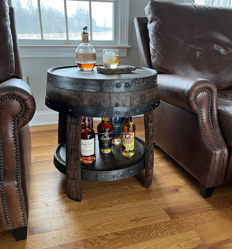 May include: A wooden barrel end table with a metal top and a lower shelf. The table has a rustic brown finish and is decorated with metal accents. There are four bottles of liquor on the shelf, including Old Forester Bourbon, Weller Special Reserve Bourbon, and a bottle of Mal&ouml;rt.