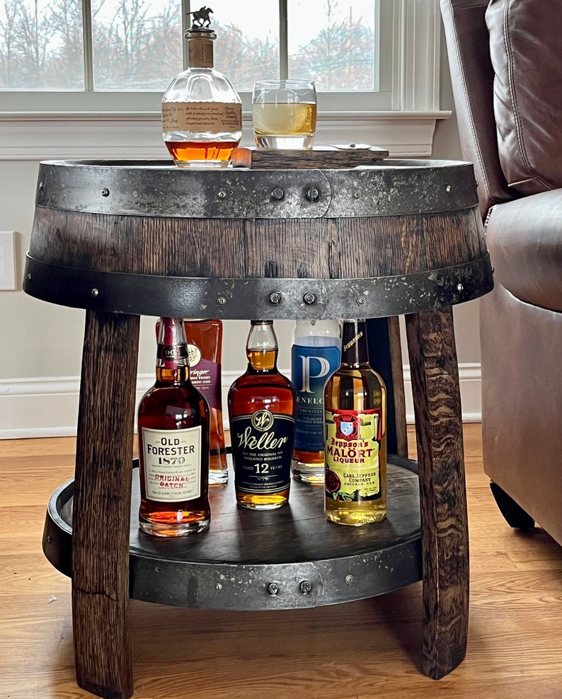 May include: A wooden barrel bar cart with a metal top and a lower shelf. The cart is filled with various bottles of liquor, including Old Forester 1870, Weller 12 Year Old, and Leopold Bros. Malort Liqueur.