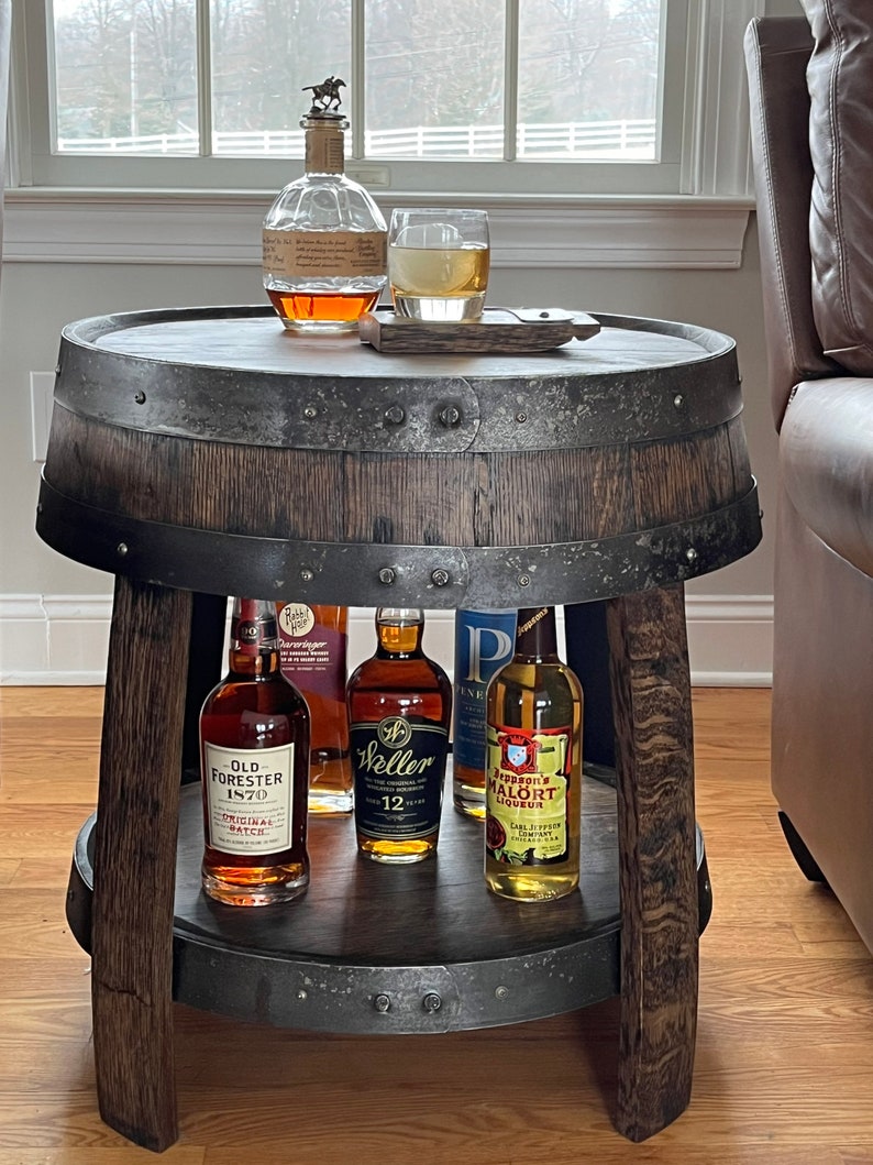 May include: A wooden barrel end table with a metal band around the top and bottom. The table has a shelf and is holding several bottles of liquor, including Old Forester 1870 Original Batch, Weller 12 Year Old, and Jeppson's Malort Liqueur.