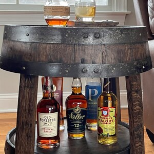 May include: A wooden barrel table with two tiers. The top tier has a glass of amber liquid and a bottle of bourbon. The bottom tier has five bottles of liquor, including Old Forester 1870 Original Batch, Weller 12 Year Old, and Jeppson's Mal&ouml;rt Liqueur.
