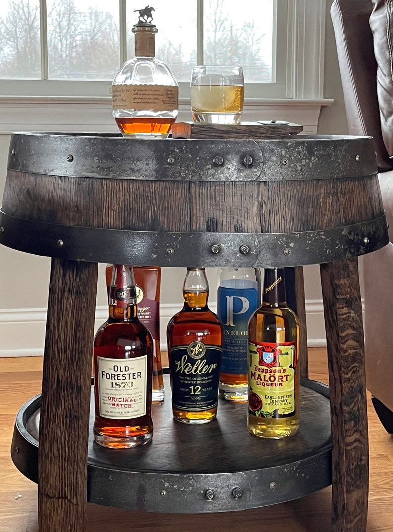 May include: A wooden barrel table with two tiers. The top tier has a glass of amber liquid and a bottle of bourbon. The bottom tier has five bottles of liquor, including Old Forester 1870 Original Batch, Weller 12 Year Old, and Jeppson's Mal&ouml;rt Liqueur.