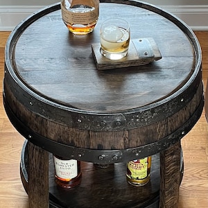 May include: A two-tiered wooden barrel end table with a dark wood finish. The top tier has a glass of whiskey and a bottle of Old Forester bourbon. The bottom tier has a bottle of Old Forester bourbon and a bottle of Amaro Averna liqueur.