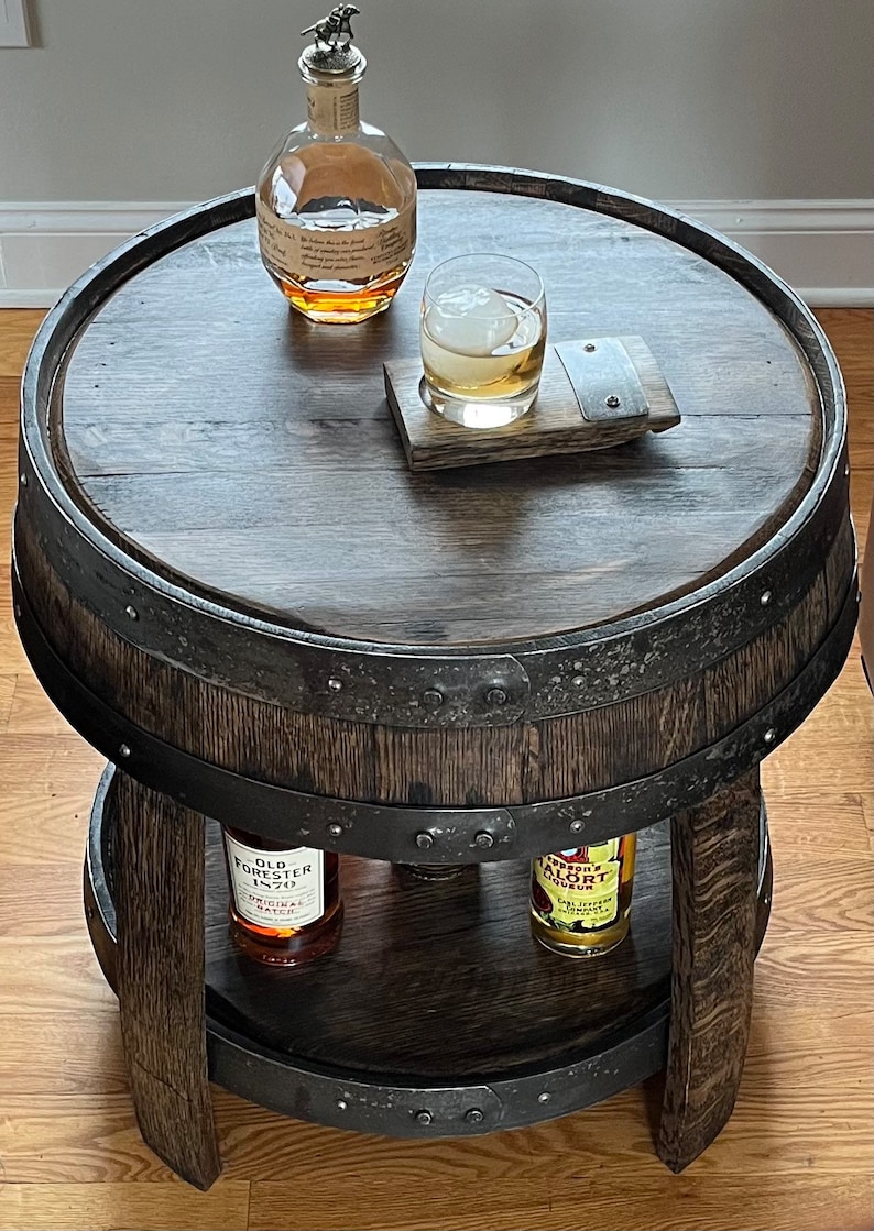 May include: A two-tiered wooden barrel end table with a dark wood finish. The top tier has a glass of whiskey and a bottle of Old Forester bourbon. The bottom tier has a bottle of Old Forester bourbon and a bottle of Amaro Averna liqueur.