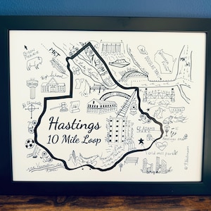 May include: Black framed art print featuring a hand-drawn map of Hastings, with the text "Hastings 10 Mile Loop". The map includes landmarks, trails, and the text "Bison Prairie".