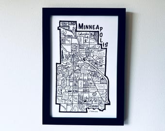 May include: A framed black and white line art map of Minneapolis, Minnesota. The illustration features hand-drawn neighborhood boundaries, street names, and landmarks with the word Minneapolis written in bold black capital letters at the top.