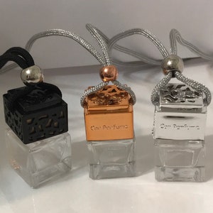 May include: Three car air fresheners with clear glass bottles and decorative tops. One has a black top, one a gold top, and one a silver top. Each has a silver or black cord for hanging. The text "Car Perfume" is visible on the gold and silver tops.