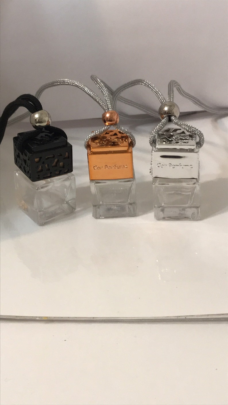 May include: Three car air fresheners with clear glass cube bottles. The lids are black, copper, and silver, with the text "Car Perfume" on the copper and silver lids. Each has a silver or black cord for hanging.