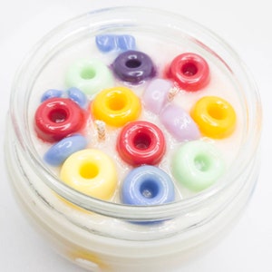 Pu&ograve; includere: Una candela in barattolo di vetro con inserti di cera colorati a forma di anello nei toni del rosso, giallo, verde, blu e viola. La candela ha due stoppini e una base di cera bianca cremosa. Sfondo bianco.